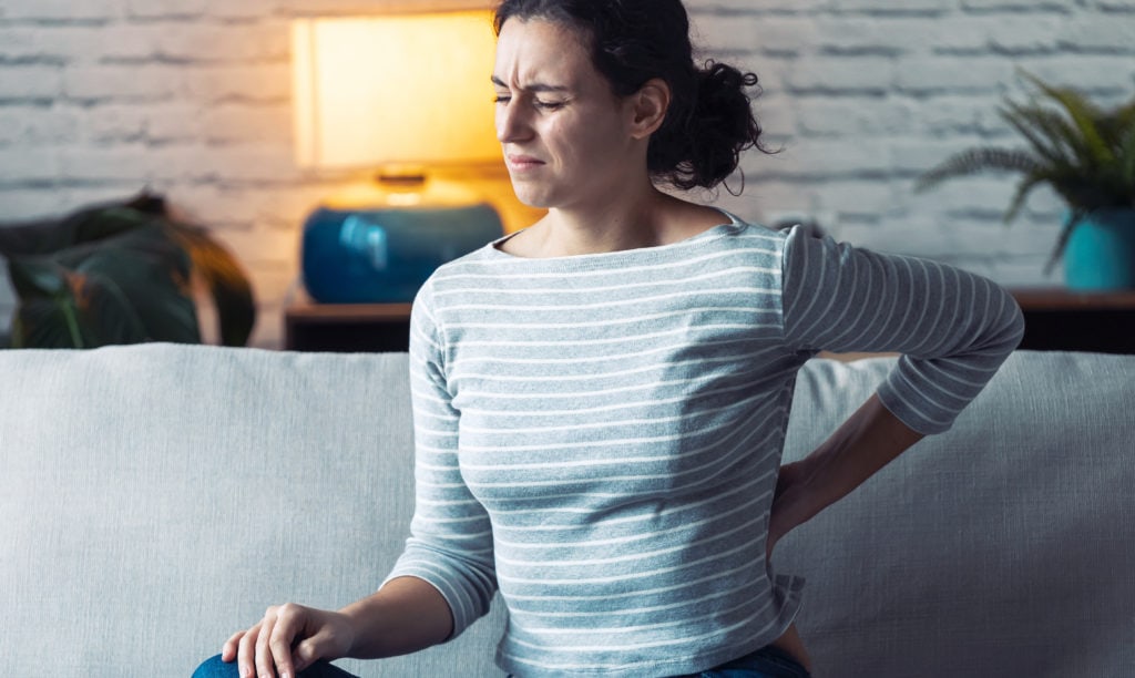 Young woman with back pain sitting on the sofa in the living room at home.